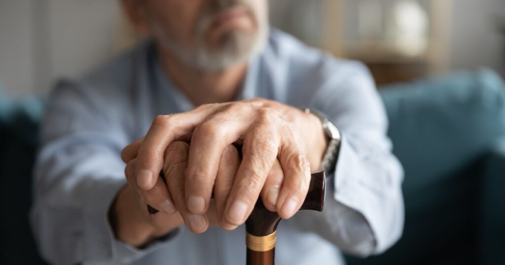Close up of old man hands hold wooden cane look in distance thinking pondering, disabled mature 60s male feel lonely abandoned lost in thoughts missing mourning, elderly solitude concept