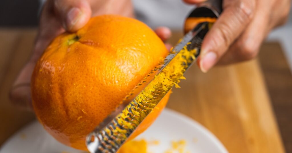 A zester zesting orange skin on a white plate on a wooden tabletop.