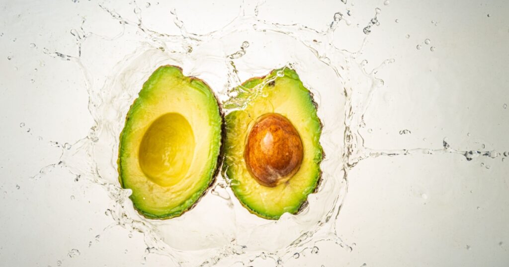 Two halves of avocado splashing into clear water, isolated on light blue background. Health food concept