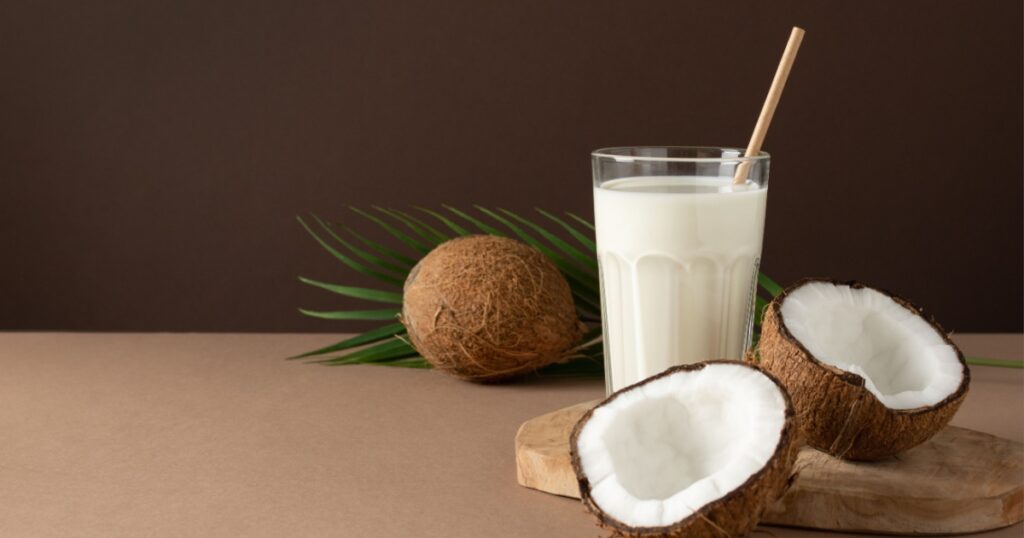 A glass of coconut vegan milk with halves of nuts over brown background.