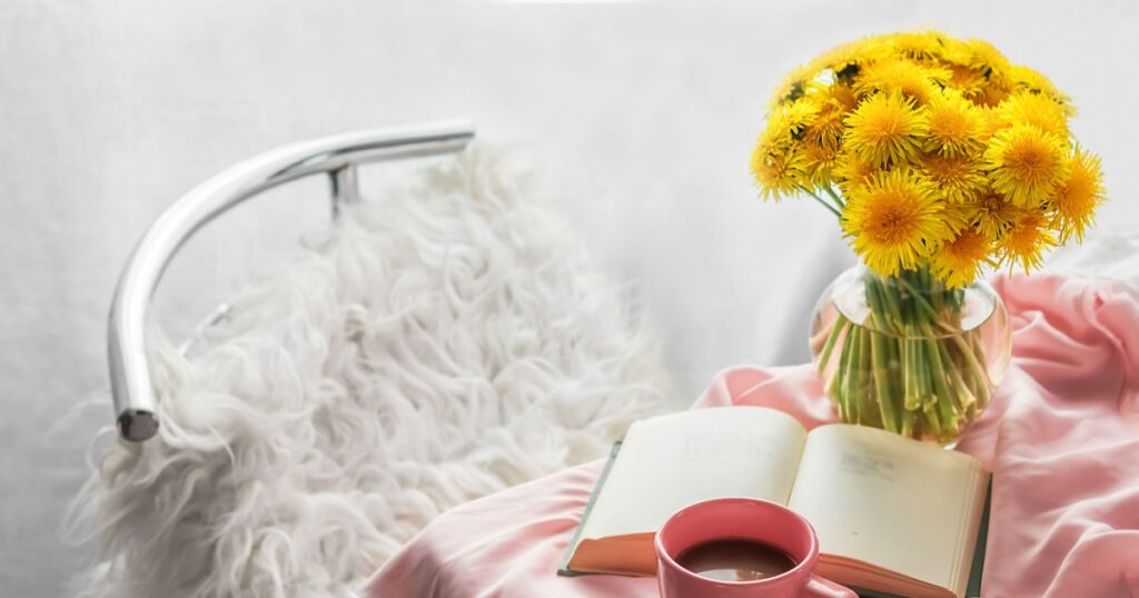 Romantic morning atmosphere Bouquet of yellow dandelions in a vase, an open book and a cup of coffee on a table on a pink silk tablecloth Concept of a cozy stylish home interior Lifestyle