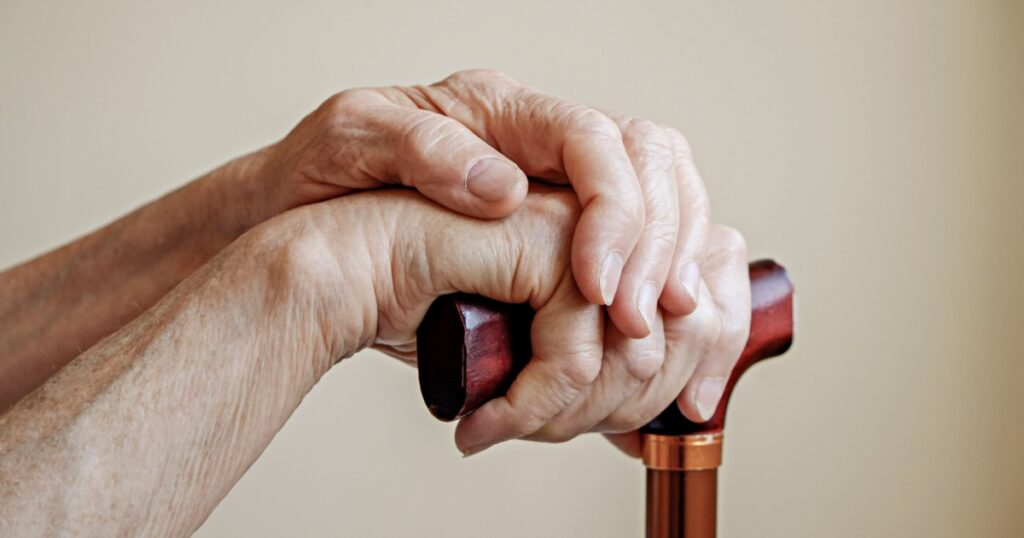 Macro shot of elderly woman's hands holding the wooden handle of a metal walking cane. Close up, copy space for text, neutral background.