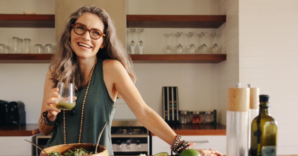 Healthy senior woman smiling while holding some green juice in her kitchen. Mature woman serving herself wholesome vegan food at home. Woman taking care of her aging body with a plant-based diet.