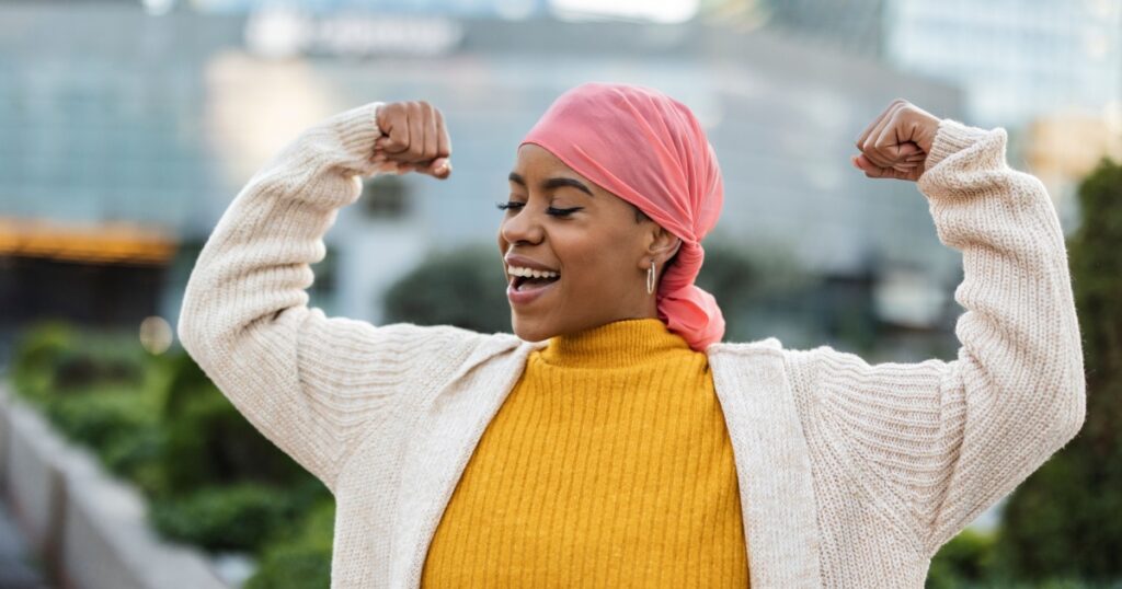 Latina woman, fighting breast cancer, wears a pink scarf, and clenches her arms as a survivor fighter
