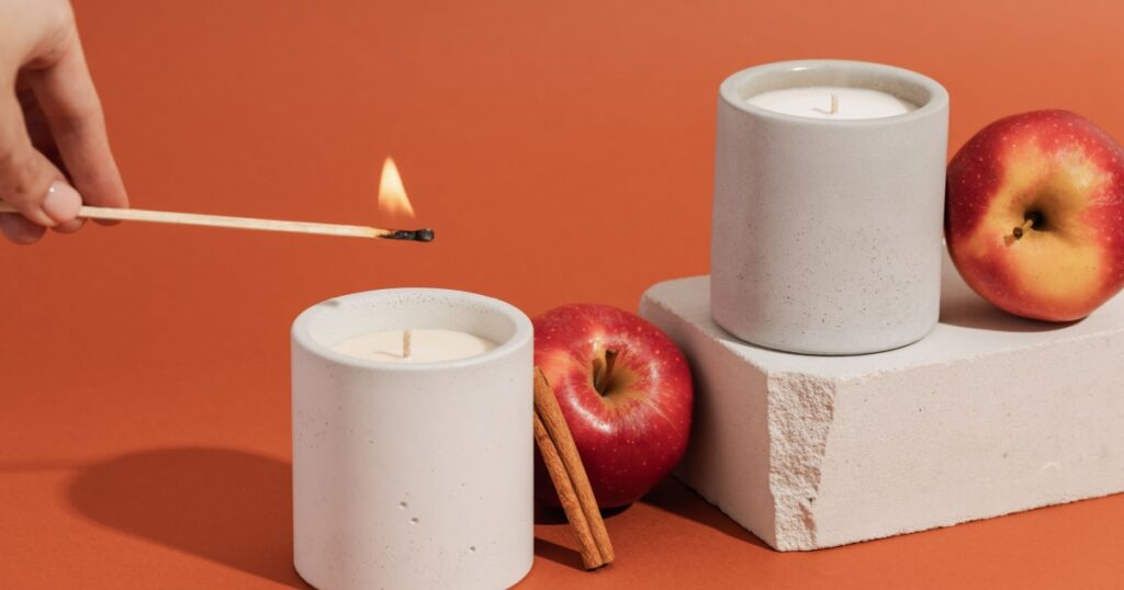 A hand lights a candle with a match stick. Aromatic soy candles in gray concrete caps with apple and cinnamon. Poster banner for candle shop, beauty, spa