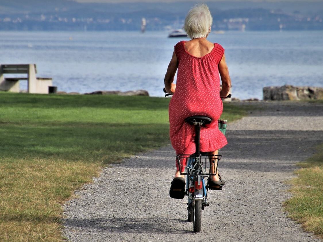 Elderly woman riding a bike. Gravel trail, grass, ocean, and mountains in the background. 