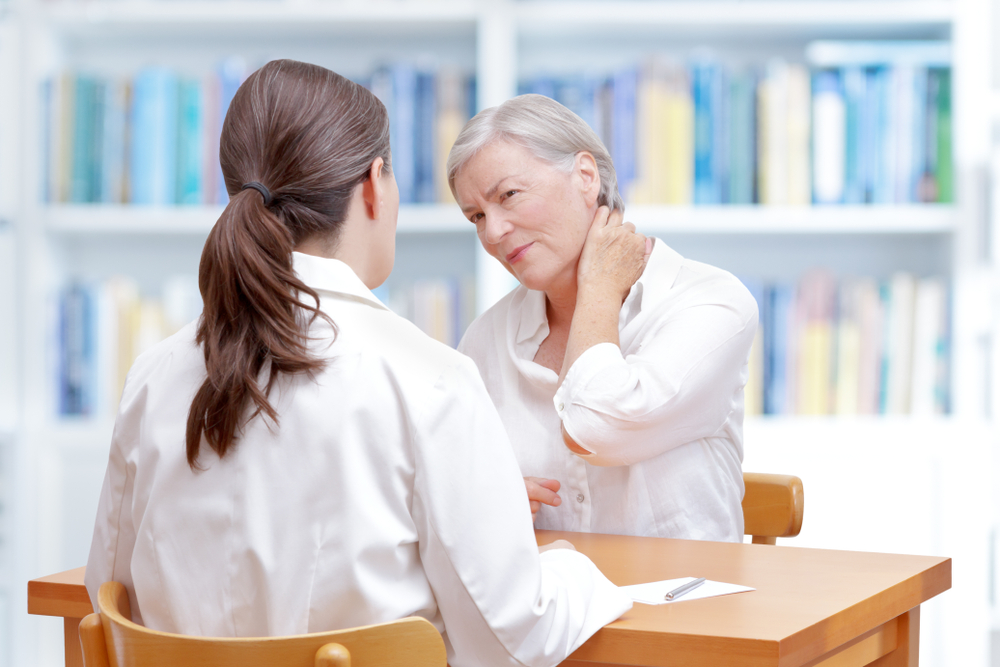 Senior female patient consulting her physician or doctor on account of chronic neck pain caused by fibromyalgia, osteoporosis or a herniated disc.
