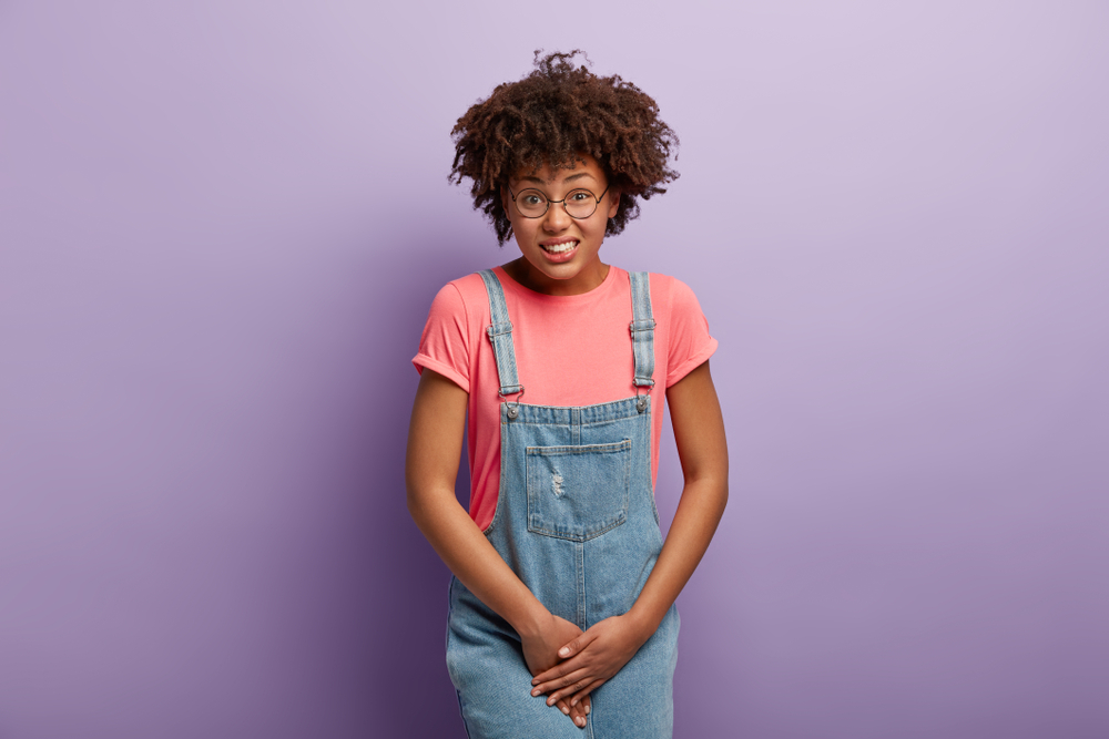 Unhappy dark skinned woman holds crotch, needs toilet, has problematic situation, wears pink t shirt and denim sarafan, suffers from cystitis, isolated over purple background. People and urgency