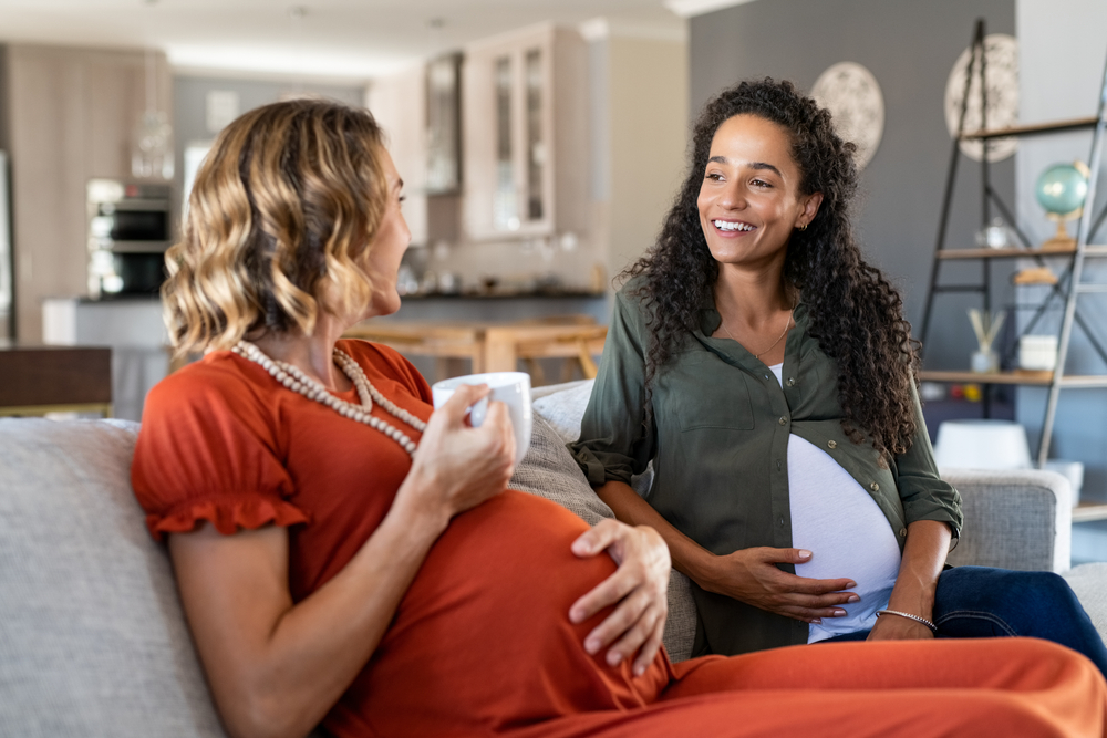 Two multiethnic pregnant women enjoying tea time at home. Smiling pregnant women talking and looking at each other. Happy girls both with a baby bump relaxing on couch and have a friendly conversation
