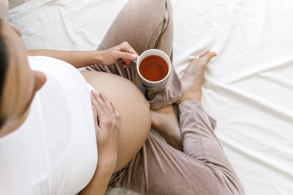 pregnant woman sitting on the white bed and holds a cup of tea in her hand in morning pregnancy, drinks, rest, people and expectation concept. Healthy nutrition for pregnant women. Pregnancy.
