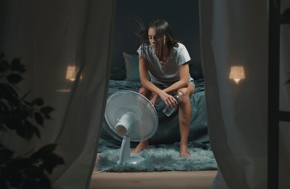Exhausted woman suffering suring the heatwave, she is holding a water bottle and sitting in front of a cooling fan in the bedroom

