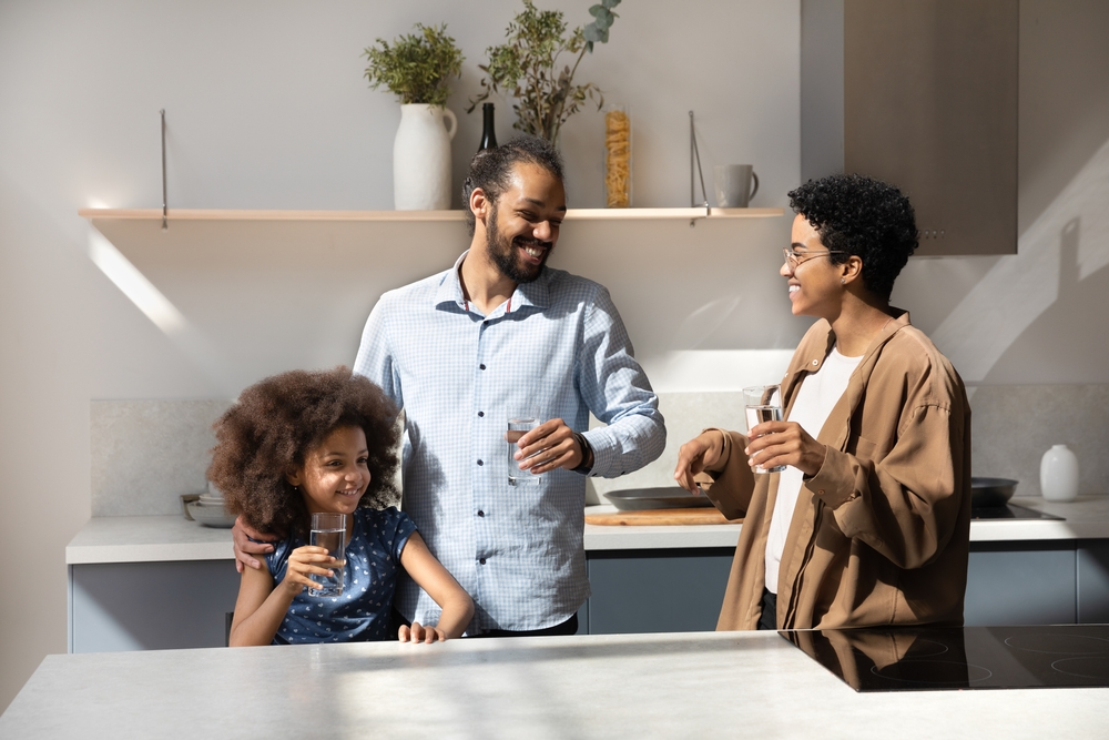 Happy laughing African American family couple and healthy daughter kid drinking clean still pure water in home kitchen, holding glasses, talking, laughing, keeping healthy hydration