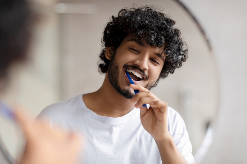 Oral care concept. Young indian man cleaning teeth with toothbrush, smiling to his reflection in mirror, doing toothcare hygiene routine in the morning in bathroom
