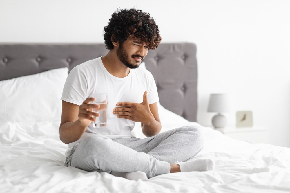 Unhappy millennial indian guy suffering from heartburn, waking up in morning, sitting on bed and holding glass of water, wearing pajamas, copy space. Acid reflux, pain in stomach concept
