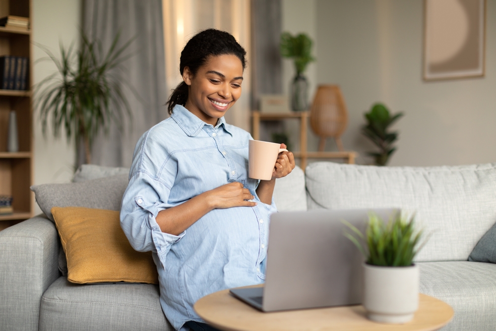 Pregnancy Lifestyle. Happy Pregnant African American Woman Using Laptop Computer Watching Movie Online And Drinking Coffee Sitting On Couch At Home. Internet Technology Concept
