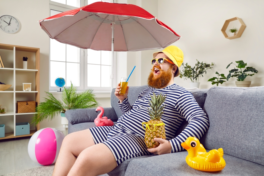 Happy bearded man in classic bathing suit sitting on sofa at home. Excited eccentric man in sunglasses and panama hat sitting under umbrella drinking cocktail enjoying summer vacation in living room
