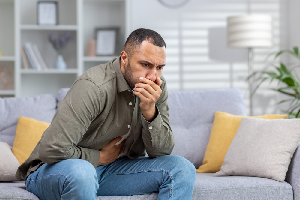 A young African American man is not feeling well. Sits at home on the couch, holds his stomach with his hand, covers his mouth with his hand, wants to vomit, feels nausea and severe pain.