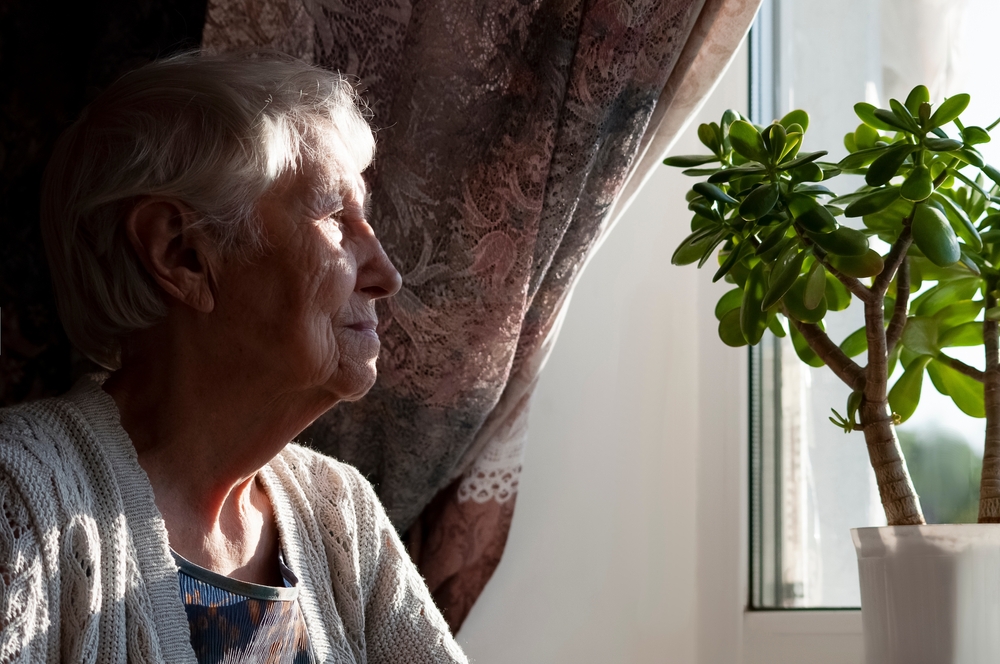 Old lonely woman sitting near the window at home
