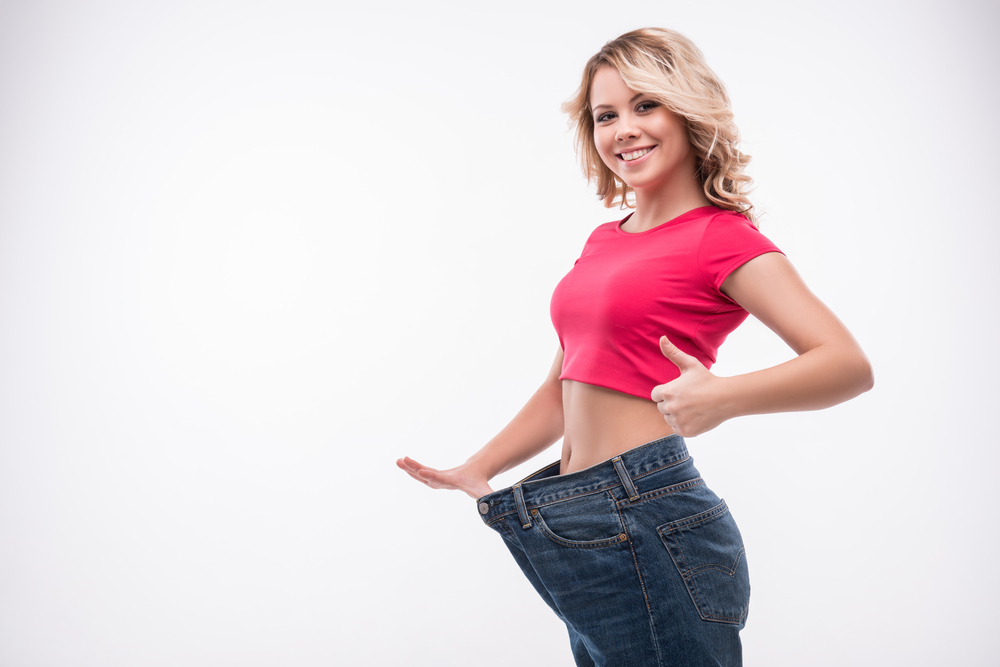 Full-length portrait of attractive slim young smiling woman in big jeans showing successful weight loss with her thumb up, isolated on white background