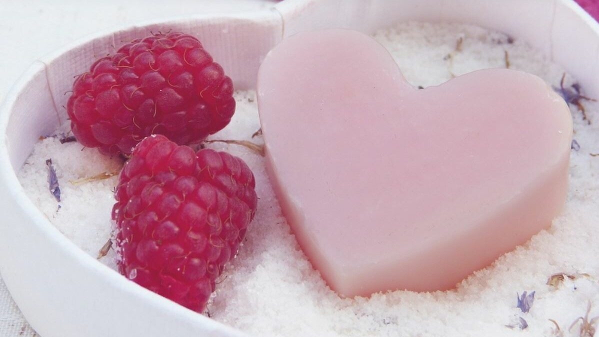 Heart shaped soap, bath salts, and raspberries in a dish. Purple flowers in the background. 
