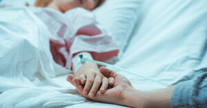 Hospice Nurse Reveals ‘Worst’ and ‘Best’ Diseases to Die From hand of patient in hospital bed placed in the hand of a visitor. End of life, hospice care concept