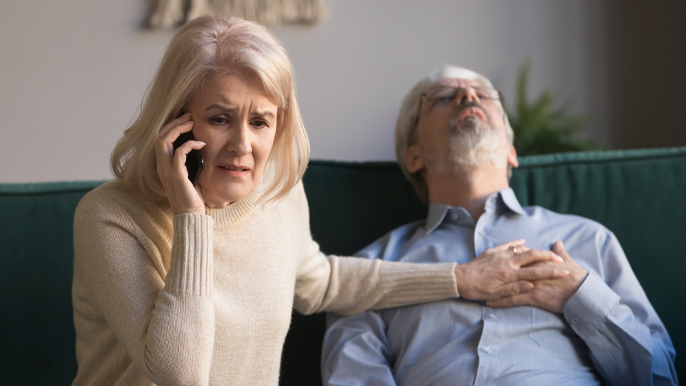 Desperate aged wife holds phone makes 911 emergency call while her sixty years husband lies on couch, man feeling crushing chest pain, loss of blood supply, shortness of breath, heart attack concept
