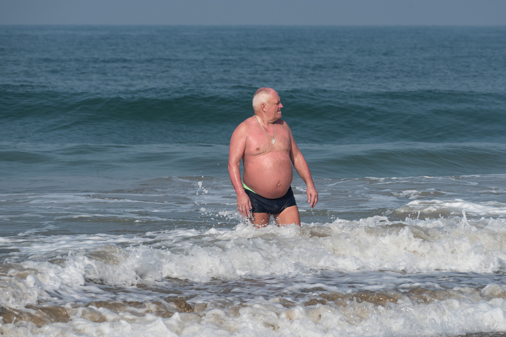 03/10/2019 - Colva, Goa, India: An old white man with large beer belly standing in shallow sea with blue ocean and sky background. An obese man in shorts at a beach coming after a swim in the sea.