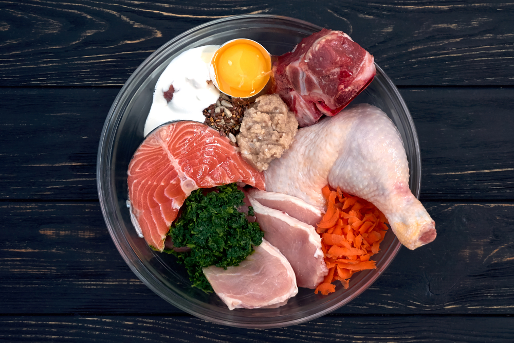 Natural raw dog food in bowl on black wooden background Meat fish eggs vegetables bones organic food supplement