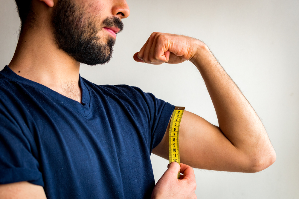 Bearded thin man measuring biceps, muscles of his left arm with a yellow tape measure. He's calm, serious, quiet. Wearing blue t-shirt. White background.