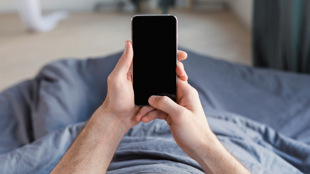 Unrecognizable Guy Using Mobile Phone With Blank Screen Lying In Bed At Home. Panorama, Mockup, POV