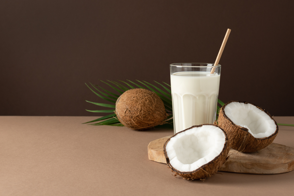 A glass of coconut vegan milk with halves of nuts over brown background.
