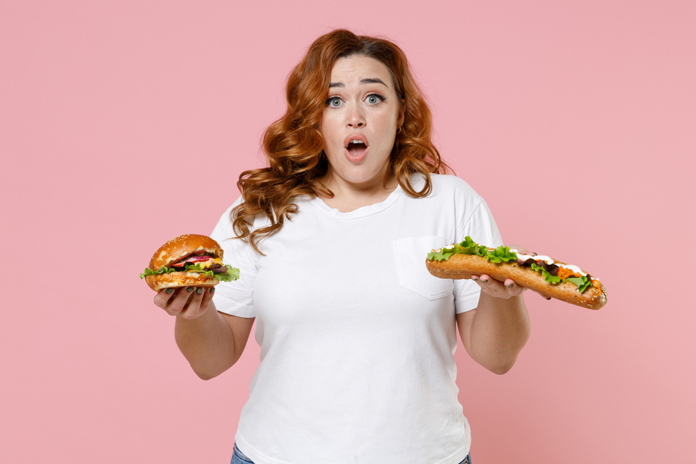 Worried young redhead plus size body positive woman 20s in white casual t-shirt hold american classic fast food hot dog burger looking camera isolated on pastel pink color background studio portrait