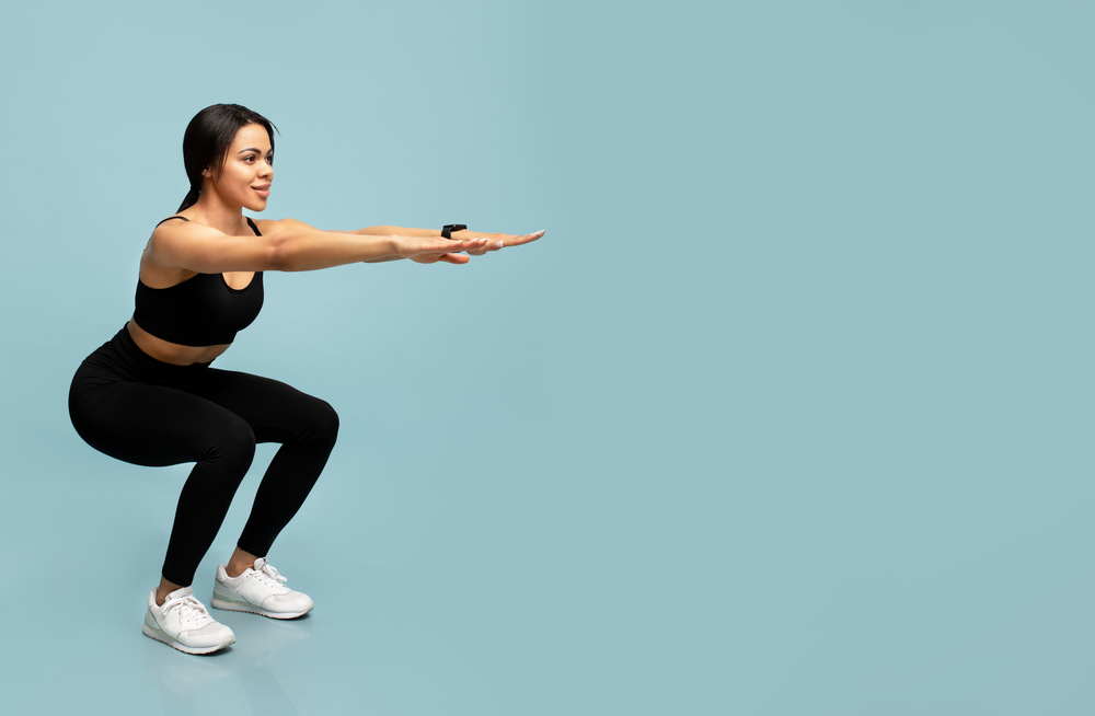 Fitness and sport. Slim african american lady in sportswear doing deep squat exercises, working out over blue studio background with empty space. Full length, banner