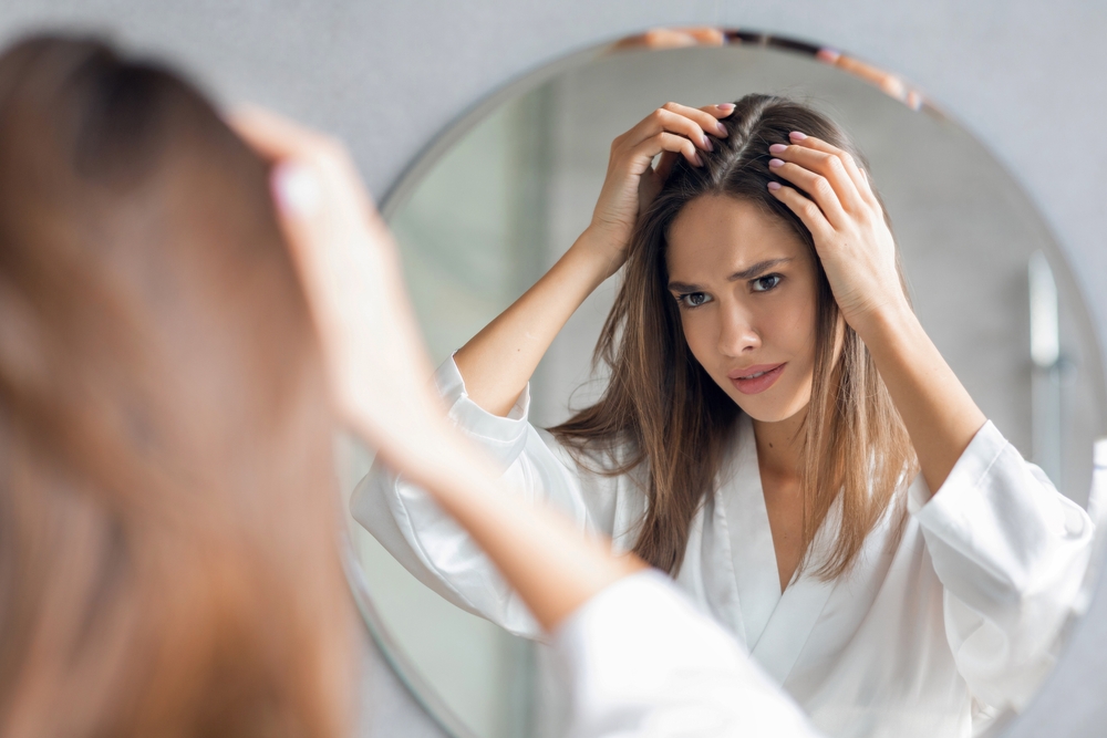 Upset Young Woman Standing Near Mirror In Bathroom And Looking At Her Hair Roots, Frustrated Millennial Lady Suffering Dandruff Or Hairloss Problem, Selective Focus On Reflection, Closeup