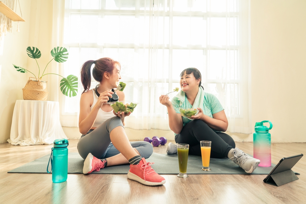 Two Asian women body size different in sport wear sitting healthy eating vegetable salad after exercise at home together. Dieting and exercise at home concept.
