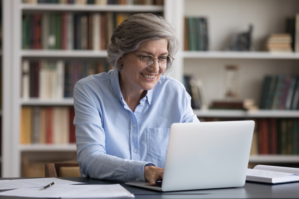 Happy successful smart old senior 60s woman in eyeglasses looking at laptop screen, working online from home, enjoying distant study, communicating in internet or web surfing information remotely.

