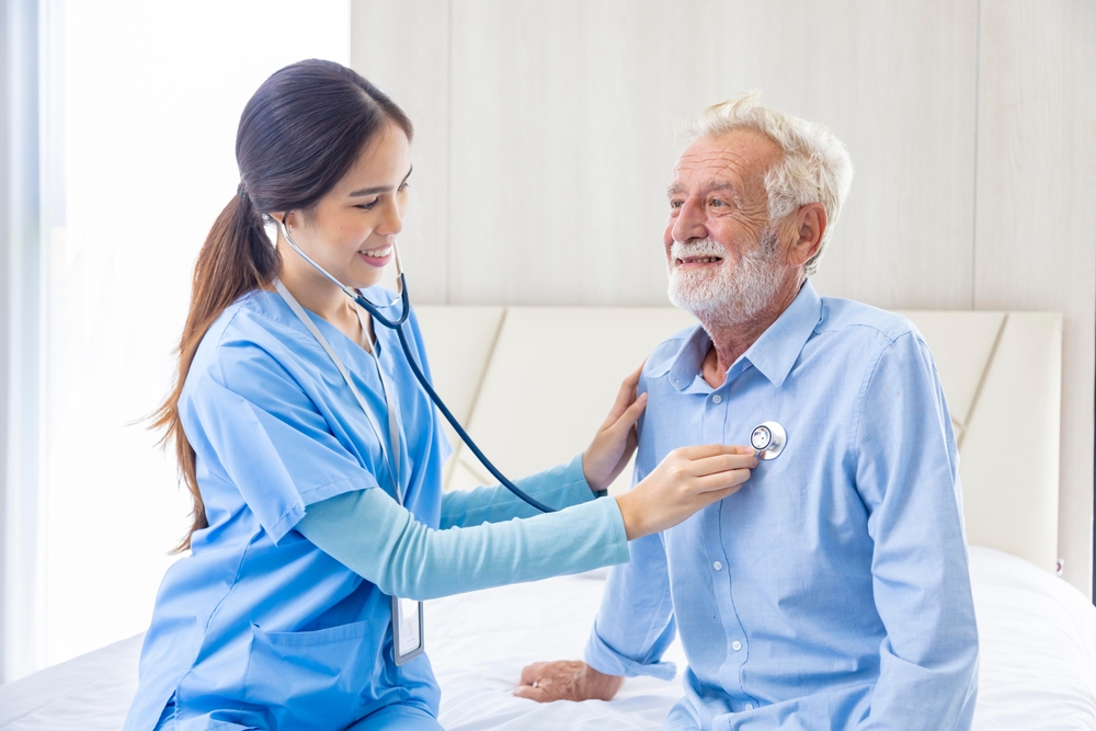 Hospice nurse or professional caregiver using stethoscope on Caucasian man in bed for diagnosing lung cancer and heart rate at pension retirement center for home care rehabilitation and post treatment