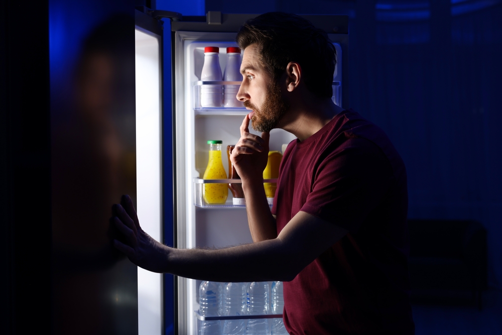 Man choosing food from refrigerator in kitchen at night. Bad habit
