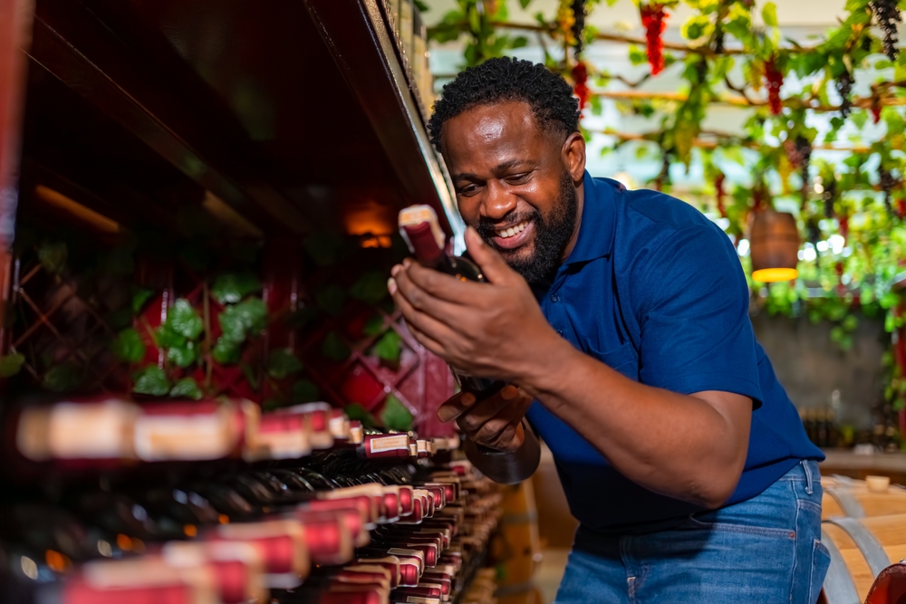 Professional African man sommelier tasting and smelling red wine in wine glass at wine cellar with wooden barrel at wine factory. Alcohol liquor shop, brewery winery industry and winemaker concept.