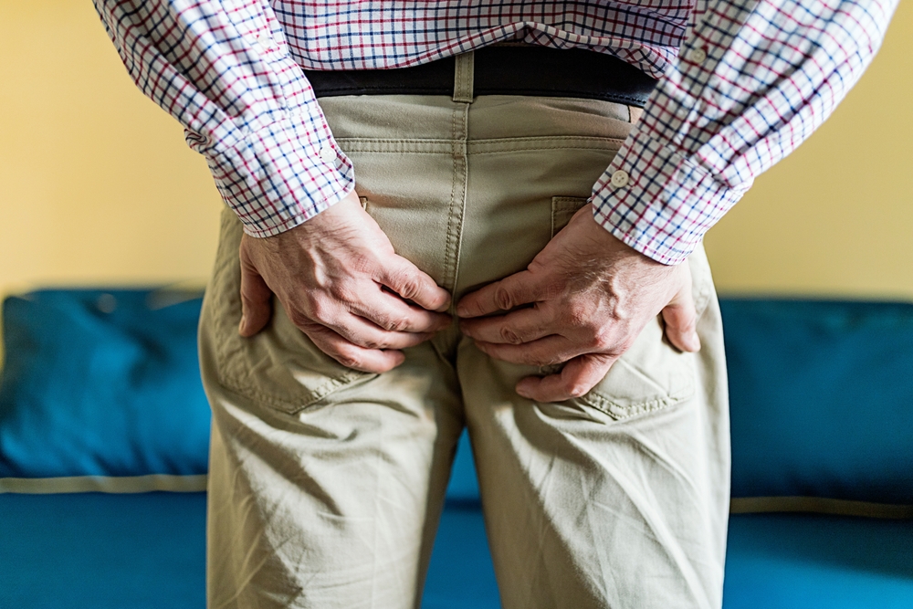 Young Man Has Pain In The Butt, Hemorrhoids, Anal Disorders. Guy With Pain Holding His Ass With His Hand. Itching In The Anus. Pain In The Rectum Due To Cracks