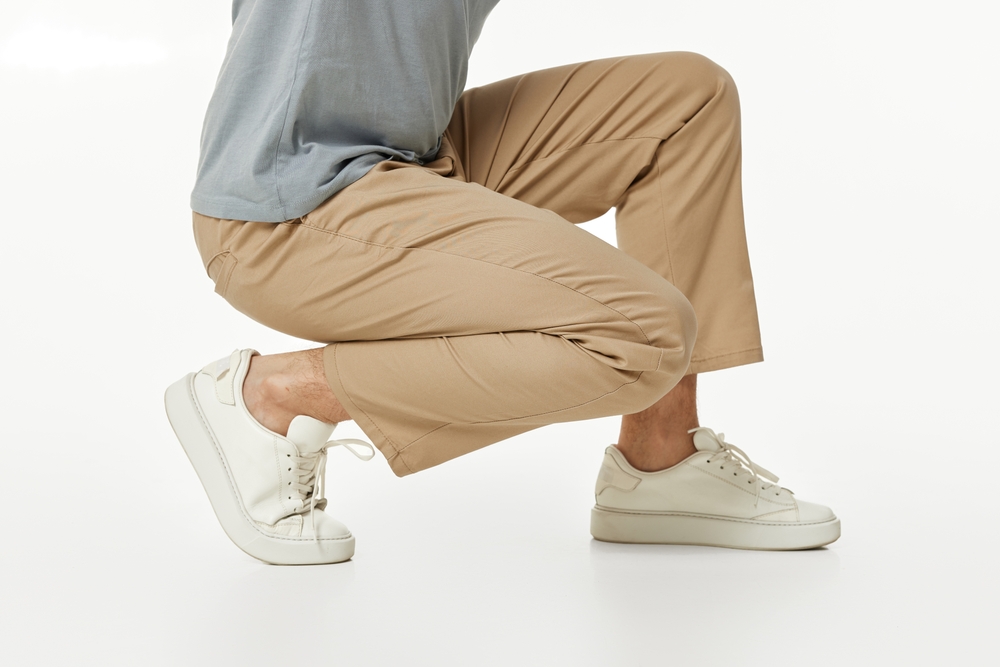 man wearing white sneakers and casual beige pants squatting on studio background. side view
