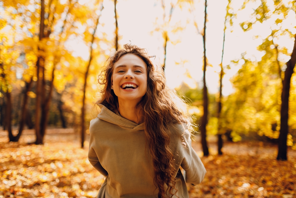 Smiling young woman enjoys the autumn weather in the forest with the yellow leaves at sunset