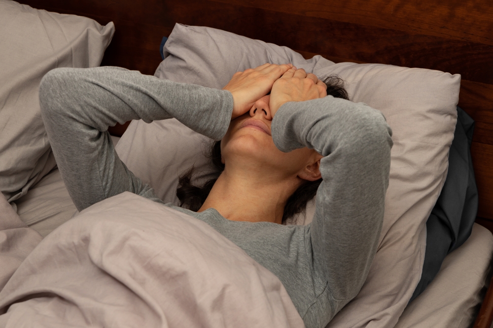 A young woman lying in bed covering her face with her hands; an ill or sick female is finding it hard to cope; a stressed or anxious lady lying in bed doesn’t want to get up
