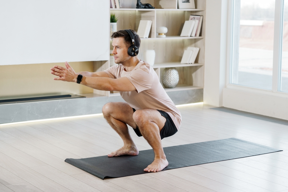 Man in headphones maintains a deep squat position, engaging in strength training on a yoga mat in a modern home.

