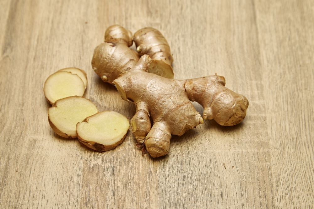 Fresh, rough-textured ginger root displayed on a wooden surface, accompanied by several slices showcasing the light yellow interior.
