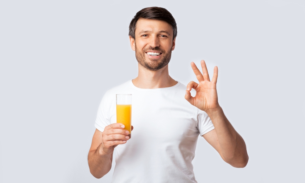 Healthy Drinks. Positive Man Holding Glass Of Orange Juice Gesturing OK Standing Over White Studio Background. Copy Space
