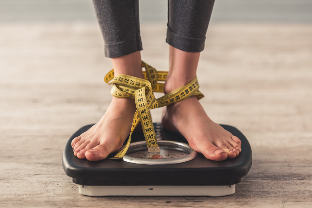 Cropped image of woman feet standing on weigh scales, on gray background. Legs winded with a tape measure
