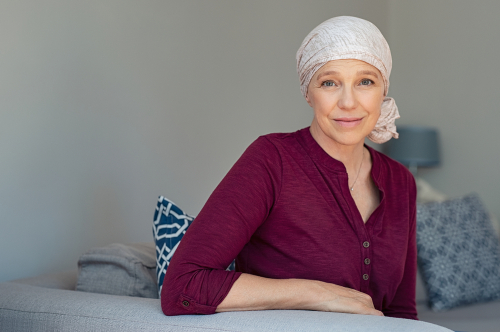 Mature woman with cancer in pink headscarf smiling sitting on couch at home. woman suffering from cancer sitting after chemotherapy sessions. Portrait of mature lady facing side-effects of hair loss.
