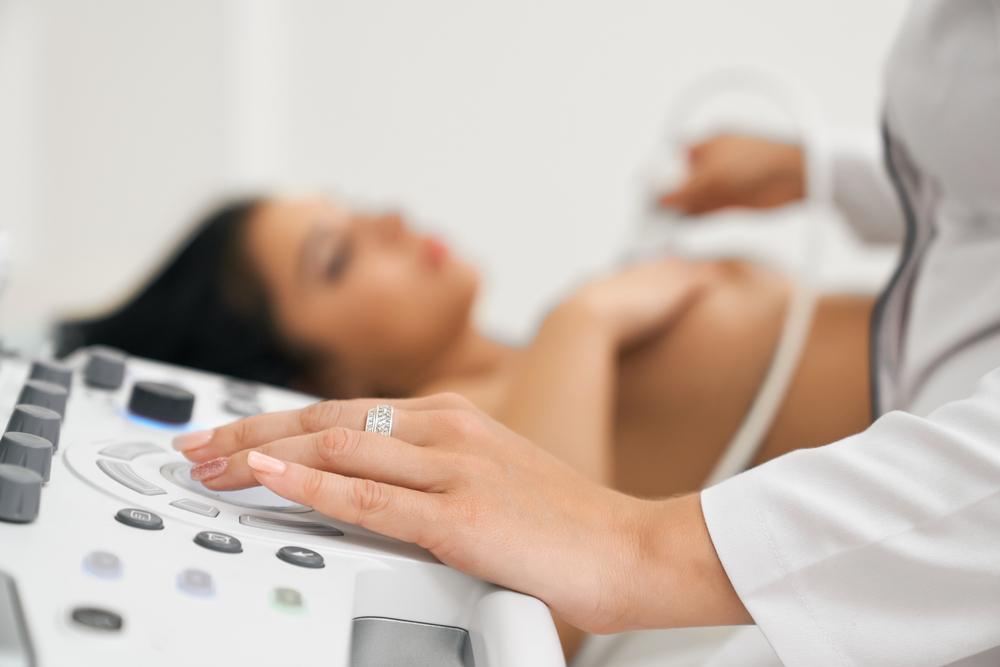 Close up of female hand with trendy manicure pushing buttons on modern ultrasound medical equipment while doing health inspection for beautiful woman with naked breast. Concept of modern medicine
