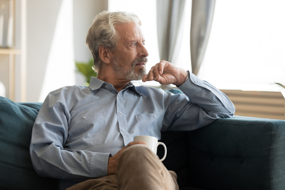 Pensive elderly 60s man sit relax on couch in living room drinking tea look in window distance thinking, thoughtful mature 50s husband rest on sofa at home feel sad pondering considering future